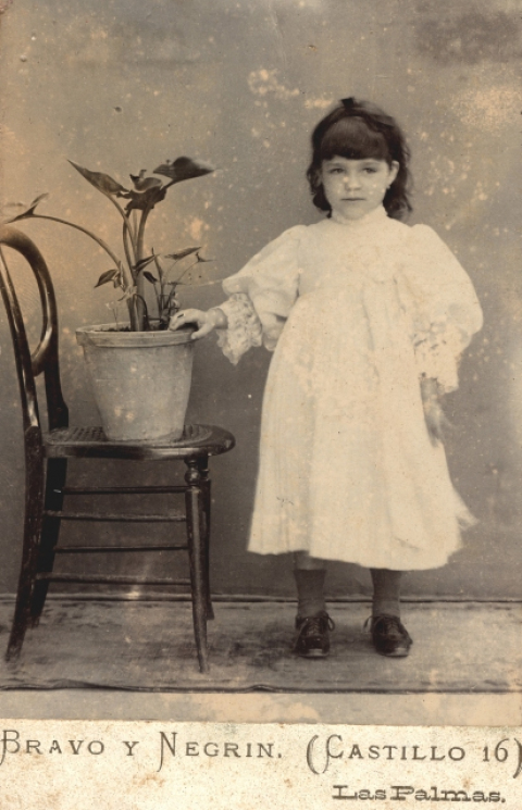 RETRATO DE NIÑA DE PIE JUNTO A SILLA CON PLANTA